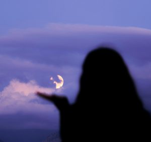 Silhouetted woman gestures toward a cloudy twilight sky with a bright moon, in Tirana, Albania.