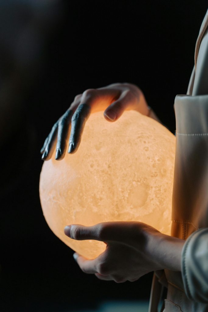A person gently holds a glowing moon lamp, casting a warm light in darkness.
